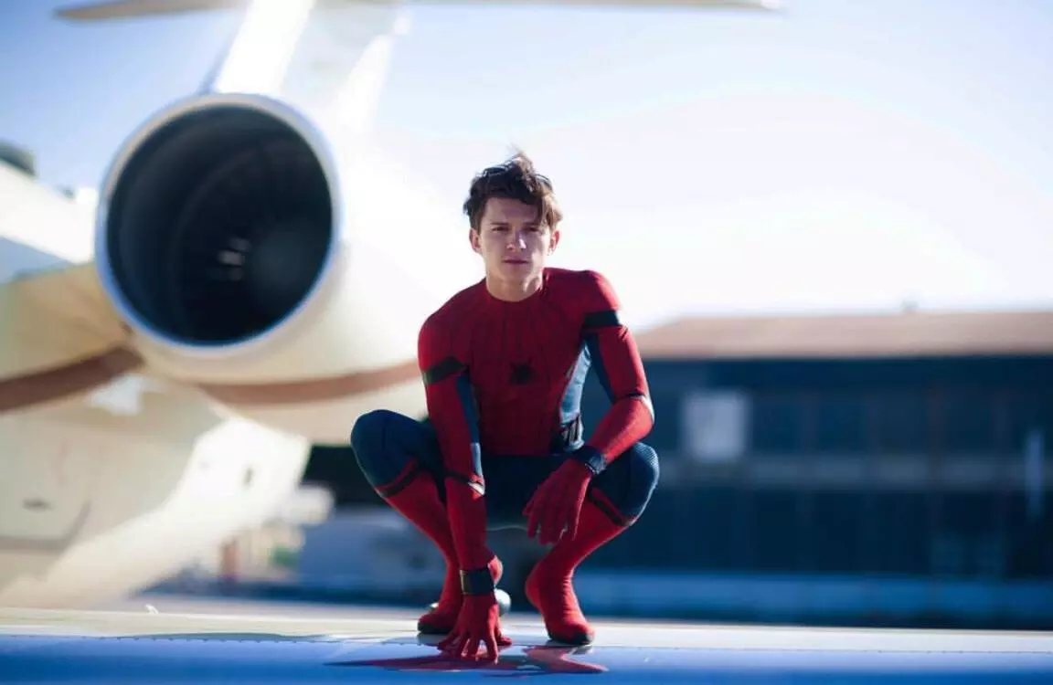 Tom Holland sebagai Spider-Man, dalam kostum merah-biru, berjongkok di dekat pesawat